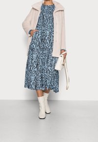 Manteau en fourrure rose clair sur une robe bleue à motifs noirs en spirale, assorti à des bottes blanches montant jusqu'aux genoux et un petit sac à main blanc.