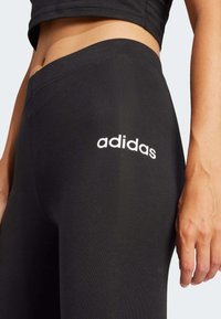 Svarta leggings gjorda av mjukt tyg med en broderad vit "adidas"-logotyp på vänster sida. Närbild av midjan och handens placering.