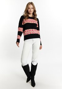Pulover colorat tricotat cu modele geometrice în nuanțe de negru, roz și galben, asortat cu blugi albi și cizme negre până la genunchi.