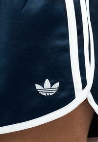 Κοντινή εικόνα σκούρων μπλε αθλητικών σορτς με λευκή επένδυση και λογότυπο Adidas trefoil στην αριστερή πλευρά πάνω από το τελείωμα.