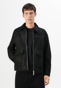 Jonge man in een zwarte zip-up jack met twee voorzakken en een kraag, staand tegen een effen witte achtergrond.