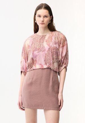 Jeune femme aux longs cheveux bruns portant des boucles d'oreilles créoles en or, un chemisier ample à motif cachemire rose et une jupe ajustée rose poudré sur fond blanc.