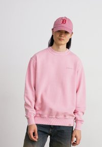 Ροζ sweatshirt με ριγωτούς καρπούς και στρίφωμα, που διαθέτει ένα μικρό λογότυπο στο στήθος. Συνδυάζεται με ένα αντίστοιχο ροζ καπέλο που εμφανίζει ένα λογότυπο.