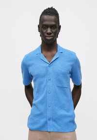 Košeľa s krátkymi rukávmi na gombíky z modrej textúrovanej látky, s golierom, predným vreckom a piatimi gombíkmi vpredu.
