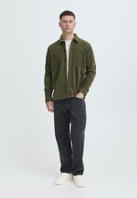 Olijfgroene, zachte fleece shirtjack met knoopsluiting, twee borstzakken, gedragen over een licht t-shirt, in combinatie met donkere jeans en witte sneakers.