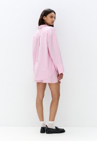 Camicia a righe rosa con bottoni, dal taglio morbido e maniche lunghe, abbinata a una gonna rosa. La modella indossa scarpe nere e calzini bianchi.