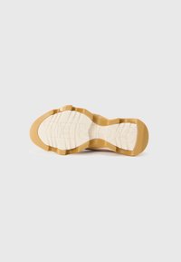 Sohle eines Schuhs mit einem wellenförmigen Design, aus hellbraunem Gummi gefertigt und mit eingeprägtem Branding. Strukturiert für Grip und Haltbarkeit.