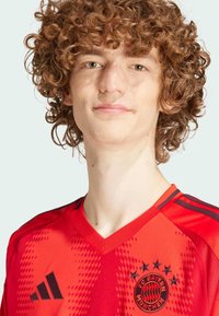 Camisa roja del FC Bayern Munich con cuello en V, logotipo negro y diseño de patrones en píxeles, hecha de un tejido ligero con detalles en negro.