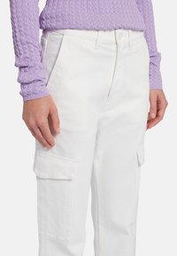 Pantaloni cargo bianchi con due grandi tasche laterali, vestibilità sartoriale e una sottile texture, abbinati a una maglietta a maniche lunghe di un leggero colore viola con texture.