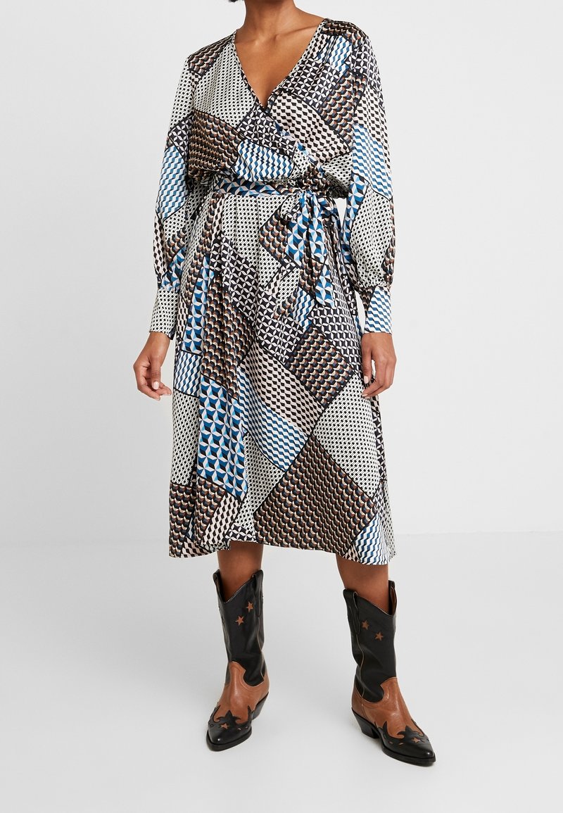 Wikkeljurk met lange mouwen, voorzien van meerkleurige geometrische prints in blauw, bruin en crème. Vastgebonden in de taille, gecombineerd met zwarte cowboylaarzen.