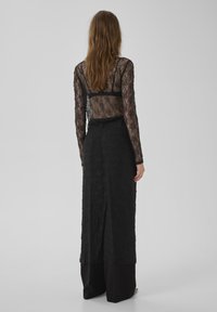 Top de encaje negro con mangas largas, superpuesto sobre una falda negra con textura floral. El conjunto se complementa con amplios pantalones negros por debajo.