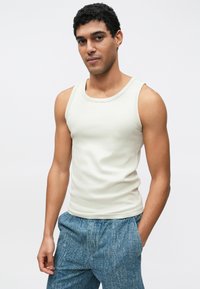 Light beige tank top with a round neckline, narrow straps, and a ribbed texture, worn with blue patterned pants. Simple and fitted design.