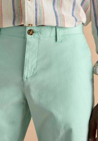 Piparmētru zaļas bikses ar gludu tekstūru, kas aprīkotas ar vienu pogu un jostām, apvienotas ar daudzkrāsainu svītrainu krekliņu augšpusē.