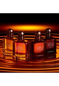 Cuatro botellas de perfume "Stronger With You" de Emporio Armani en tono ámbar con tapas redondas sobre una superficie reflectante con anillos de luz naranja.