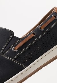 La scarpa nera in suede senza lacci presenta dettagli in pelle e perforazioni. Suola in gomma bianca con cuciture visibili lungo il bordo.