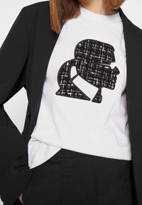 Camiseta blanca con un logo negro texturizado en forma de perfil, llevada debajo de un blazer negro ajustado, que presenta una tela suave y un corte a medida.