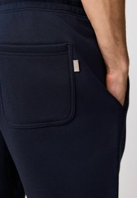 Pantalones de chándal azul marino fabricados con un tejido suave que cuentan con un prominente bolsillo trasero de diseño rectangular y una pequeña etiqueta de metal con el logo.