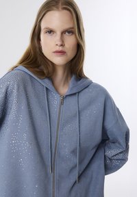 Felpa leggera di colore azzurro realizzata in tessuto morbido, con chiusura a zip e paillettes trasparenti decorative su un lato. Inclusi cappuccio e stringhe.