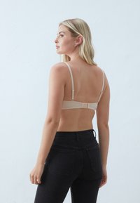 Beige vezeték nélküli melltartó állítható pántokkal, puha textúrával és hátul kapoccsal záródik. Magas derékú fekete farmernadrággal párosítva.