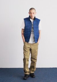Vesta pufă albastră peste un tricou alb cu grafică, asociată cu pantaloni khaki și pantofi negri. Textură netedă, croi casual și stil stratificat.