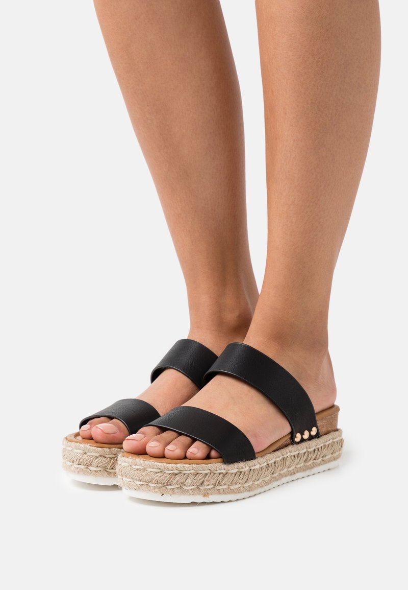 Pieds portant des sandales à plateforme noires à deux lanières avec semelles en corde tissée et petits clous dorés sur fond blanc.