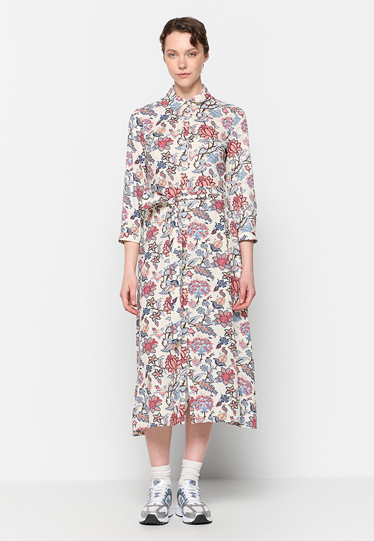 Blumiges Hemdkleid in Creme mit blauen und pinken Drucken, langen Ärmeln, Taillenschnürung und Midi-Länge, kombiniert mit Sportschuhen.