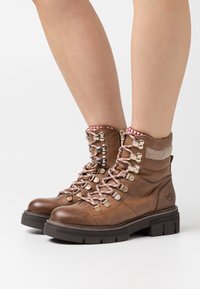 Hnědé kotníkové boty z kůže, s růžovými a bílými pruhovanými tkaničkami, stříbrnými očky a texturovaným límcem. Robustní černá gumová podrážka.