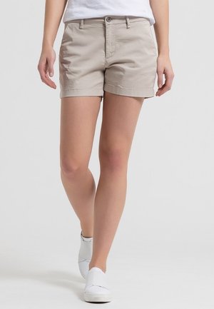 Kvinde iført beige casual shorts, hvide sneakers og en hvid top, stående med det ene ben let fremad mod en ensfarvet baggrund.