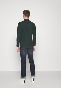 Camisa de manga larga verde oscuro con cuello abotonado, dobladillo curvado, combinada con jeans ajustados azules y zapatillas blancas. Diseño sencillo y moderno.
