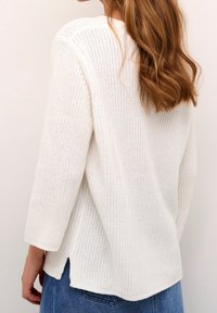 Femme aux longs cheveux ondulés portant un pull en maille blanc à manches trois-quarts et un jean en denim bleu, vue de dos.