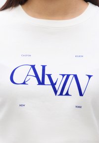 Λευκό μπλουζάκι με έντονο μπλε κείμενο που γράφει «CALVIN KLEIN NEW YORK», με ποικιλία μεγεθών και στυλ γραμματοσειράς για οπτικό ενδιαφέρον.
