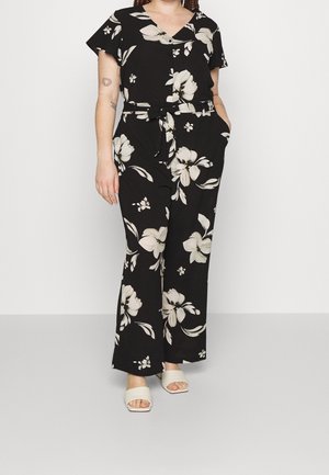 Femme portant une combinaison noire à manches courtes avec un grand imprimé floral blanc, taille ceinturée, poches et sandales blanches ouvertes à l'avant.