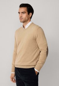 Bež jastučić s V-izrezom i dugim rukavima, s rebrastim manžetama i pojasom. Sadrži svilen patcheve na laktovima. Nosite ga preko bijele košulje s kragnom.