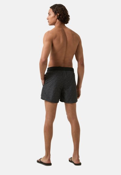Zwarte zwemshorts met een elastische tailleband, voorzien van een geometrisch patroon. Het model staat blootsvoets met de rug naar de camera.