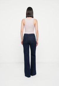Jeans evazati de culoare albastru închis cu talie înaltă și detalii vizibile de cusătură; asortați cu un top din bumbac roz deschis, văzuți din spate.