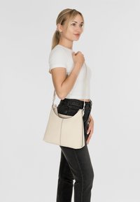 Borsa a mano in pelle beige con una texture liscia, caratterizzata da un design curvo e una tracolla regolabile, posizionata su uno sfondo grigio chiaro.
