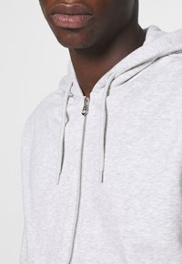Sweat à capuche gris à zip avec une finition texturée, doté d'une capuche à cordon de serrage et d'une fermeture éclair en métal. Design minimaliste sans motifs supplémentaires.