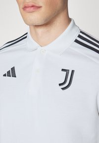 Hvit polo skjorte med krage, tre knapper, svarte striper på skuldrene, med adidas-logo og Juventus-emblem i svart broderi.