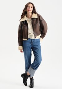Brązowa kurtka bombowiec z owczej wełny z kremowym kołnierzem, połączona z paskowanym swetrem i niebieskimi dżinsami z podwiniętymi nogawkami. Czarna para botków za kostkę dopełnia całość.