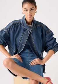 Νεαρή γυναίκα καθισμένη με σταυρωμένα πόδια, φοράει υπερμεγέθη τζιν μπουφάν, τζιν φόρεμα, χρυσούς κρίκους στα αυτιά και κόκκινες γόβες σε λευκό φόντο.