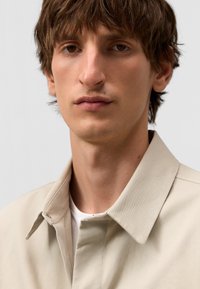 Camicia di cotone beige con colletto a punta, caratterizzata da una trama liscia e dettagli con bottoni. Maglietta bianca visibile sotto il colletto.