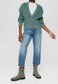 Miętowozielony sweter warkoczowy z dekoltem w serek, zapięciem na guziki i teksturowanym wzorem, w zestawieniu z jasnoniebieskimi dżinsami i szarymi botkami za kostkę.
