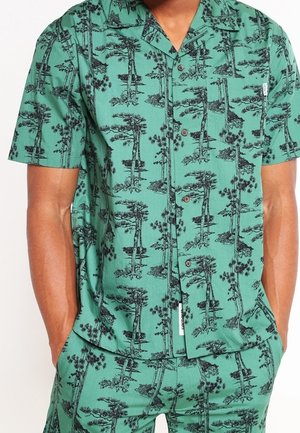 Man wearing green short-sleeve shirt and shorts set with black tree print, hands in pockets, buttoned shirt front, standing against plain background.