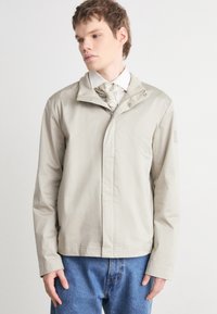 Beige zip-jakke med høy krage, lange ermer og rett snitt. Har en jevn tekstur og minimal detaljering. Kombinert med blå jeans.