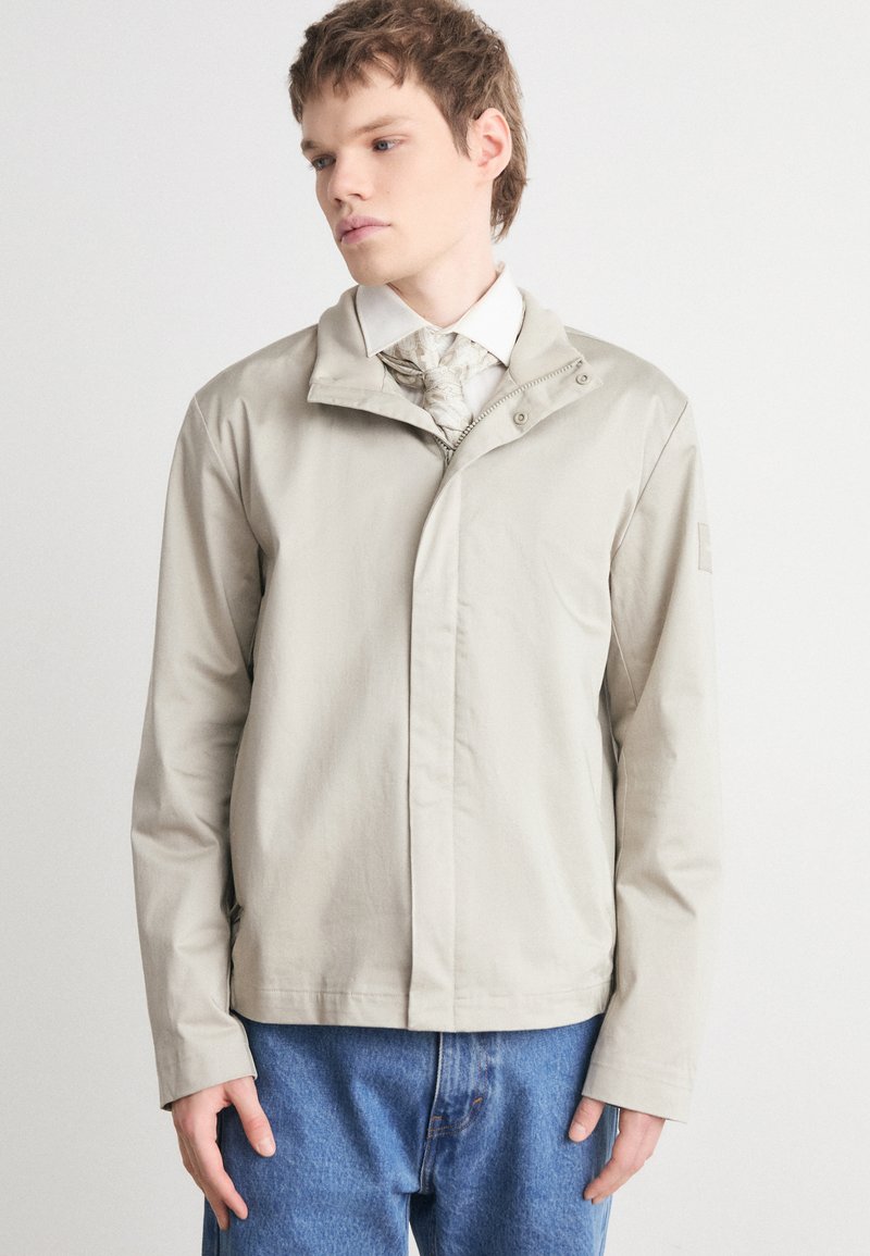 Beige zip-jakke med høy krage, lange ermer og rett snitt. Har en jevn tekstur og minimal detaljering. Kombinert med blå jeans.