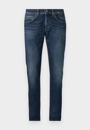 Uomo con maglietta nera, jeans blu scuro e sandali neri, in piedi con una mano in tasca su sfondo bianco semplice.
