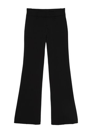 Femme aux cheveux bouclés portant un haut en dentelle noire à manches longues et un pantalon noir ajusté, debout contre un mur bleu clair uni.