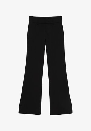 Femme aux cheveux bouclés portant un haut en dentelle noire à manches longues et un pantalon noir ajusté, debout contre un mur bleu clair uni.