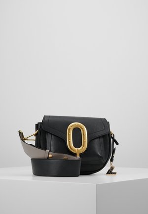 Sac bandoulière en cuir noir avec une grande boucle ovale dorée, une large bandoulière amovible et un petit pendentif doré sur une surface d'exposition blanche.