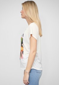 Hvid t-shirt med kort ærme og et farverigt frugtmotiv på fronten; parret med lyseblå jeans. Glat tekstur, afslappet pasform.
